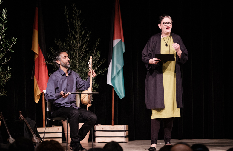 The photo shows the Brühl singer Maria Jonas and the Iraqi musician Bassem Hawar on stage on the opening evening. She is on the right side of the picture, he is sitting on the left side, framed by the German and Palestinian flag. In his left hand and on his left knee he holds his musical instrument, a knee-fiddle. In his right hand he holds the bow.